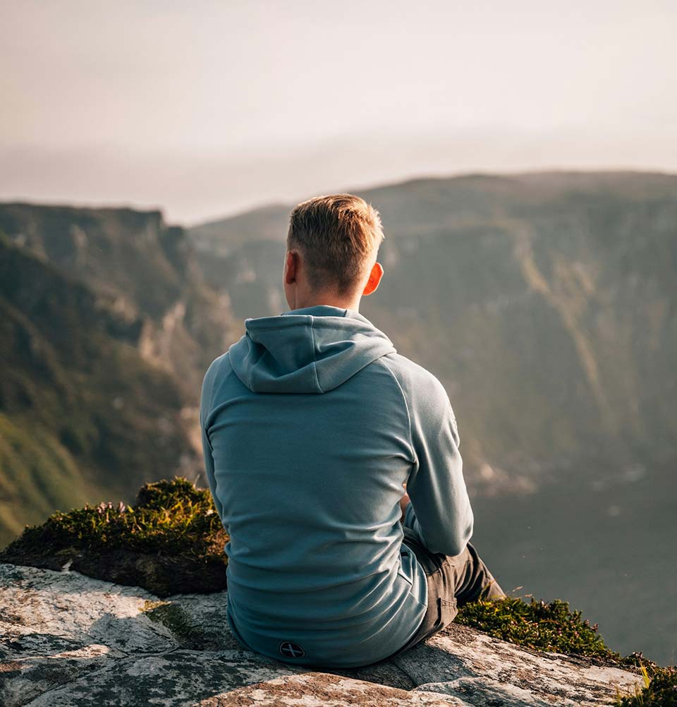 Man in a blue hoodie sitting on a cliff edge looking at the mountains.