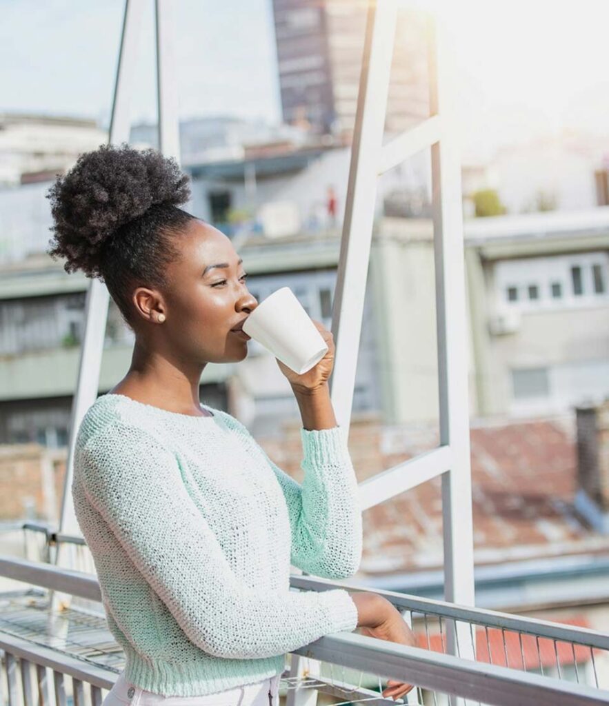 Woman in a mint sweater drinking from a mug on a sunny city balcony.