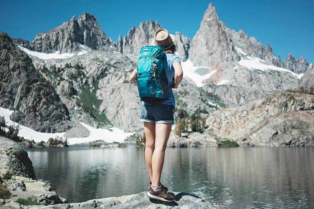 A hiker with a turquoise backpack and a straw hat stands on a rock