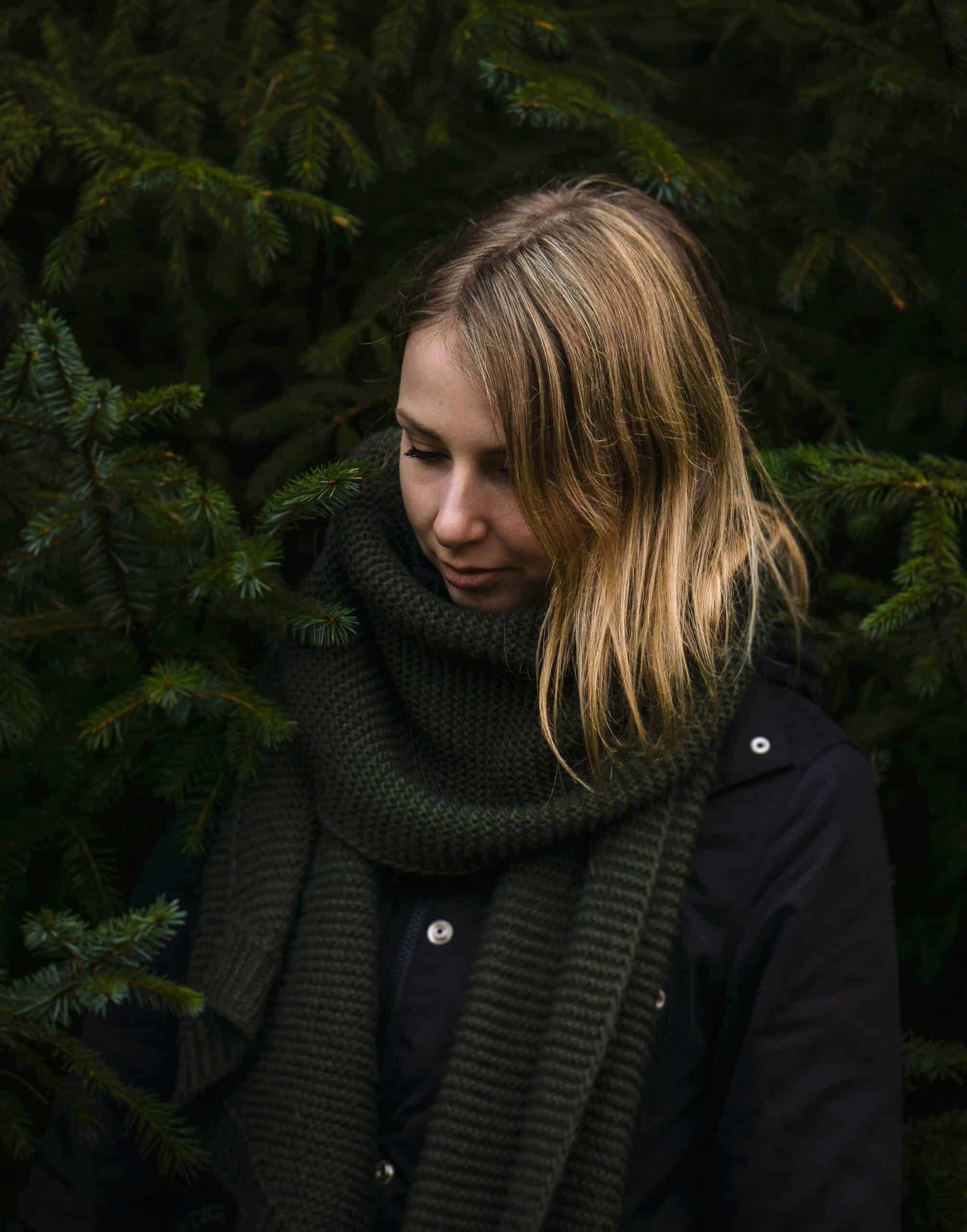 A woman standing in a forest wearing a jacket and scarf, looking down thoughtfully in a quiet reflective moment.