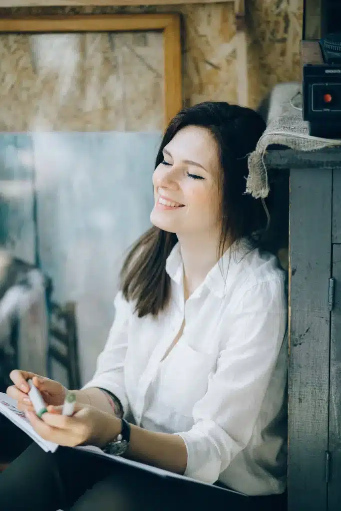 Smiling woman with dark hair wearing a white shirt holding markers and a sketchbook inside an art studio.