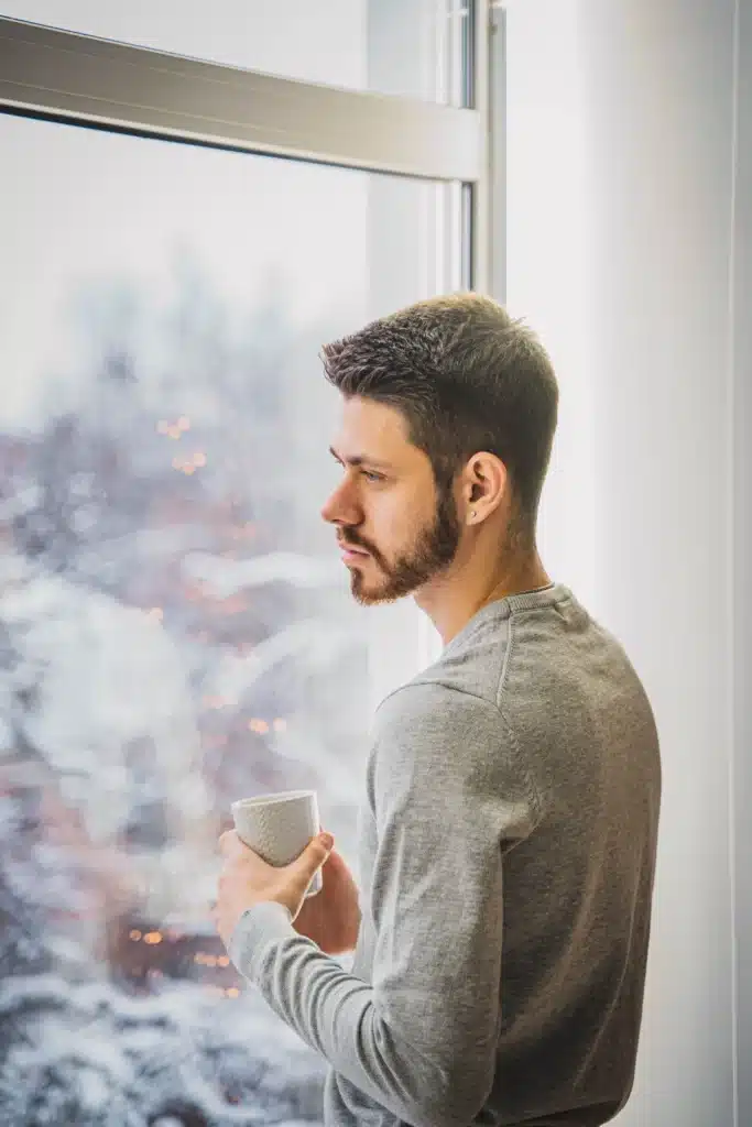 Pensive man with a short beard wearing a gray sweater holding a white mug while looking out a window at snowy trees