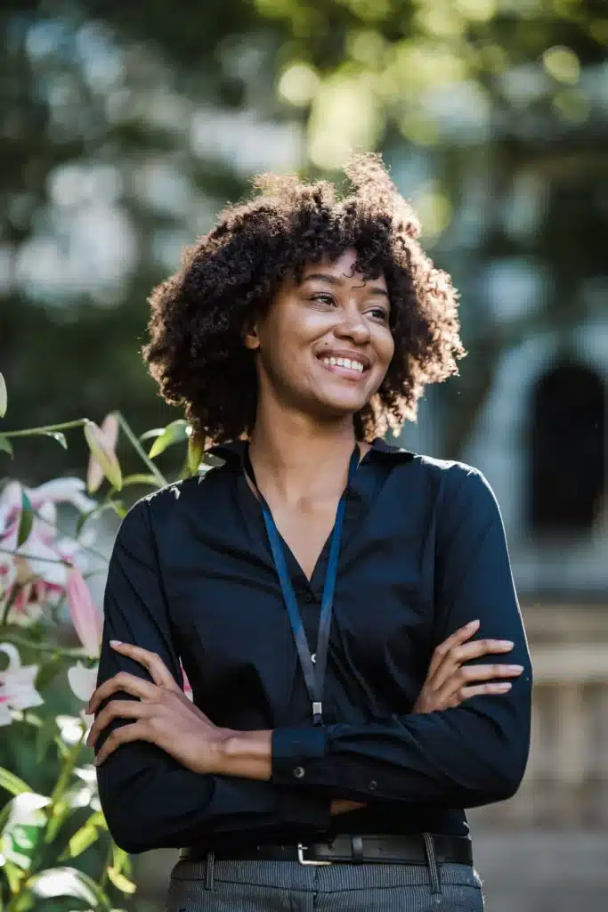 Smiling Black woman with curly hair wearing a black shirt and lanyard standing outdoors with crossed arms.