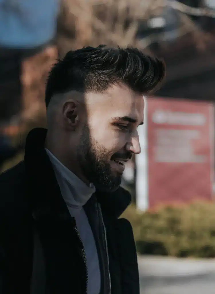 Side profile of a smiling young man with a trimmed beard and stylish fade haircut, wearing a black jacket over a white turtleneck.
