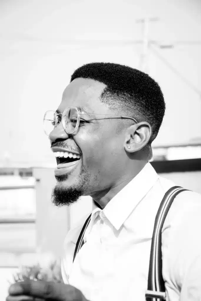 Black and white side profile portrait of a smiling Black man wearing round glasses, a button-down shirt, and suspenders, laughing cheerfully.