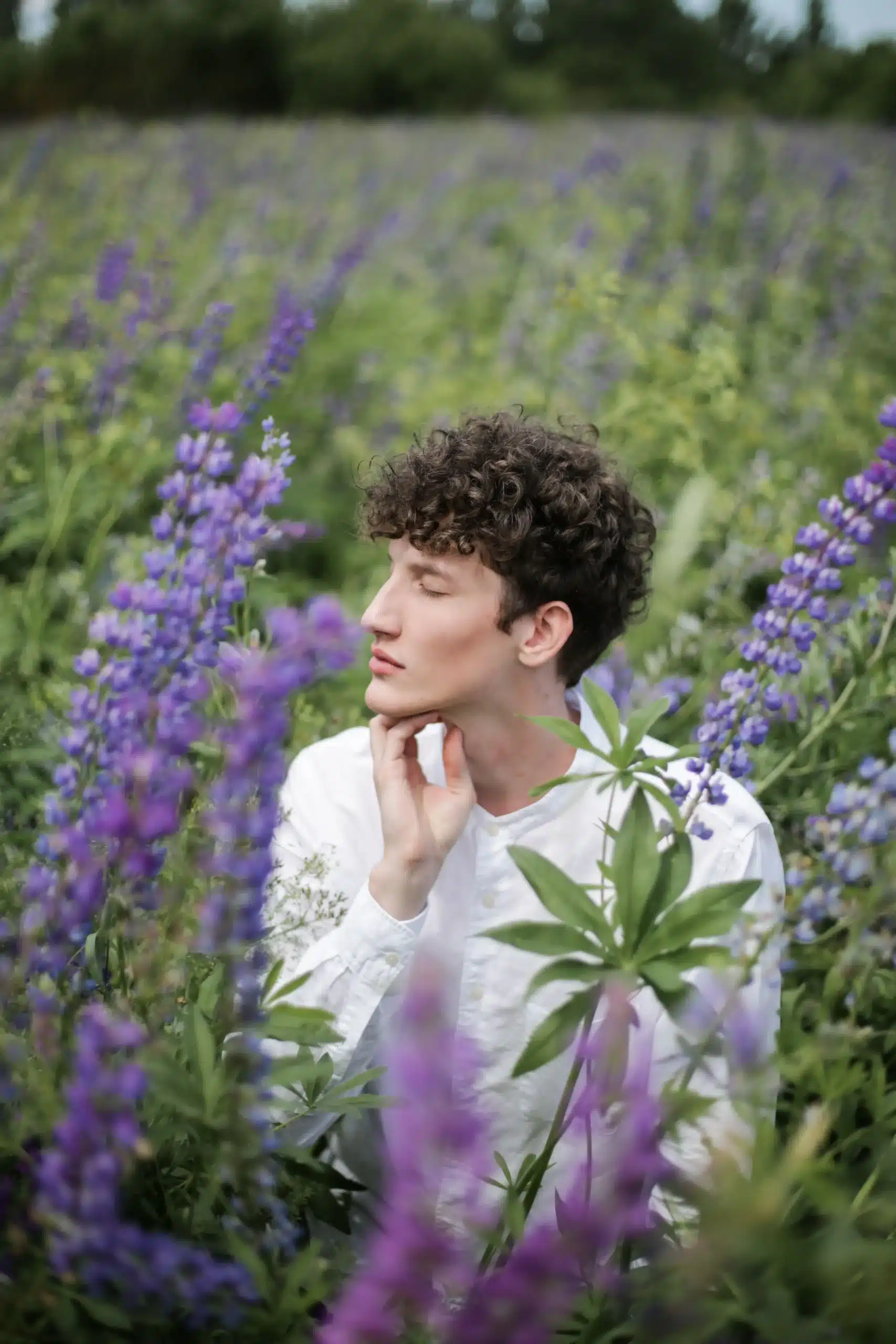 Side profile of a young man with curly hair wearing a white shirt, sitting with eyes closed in a field of blooming purple lupine flowers.