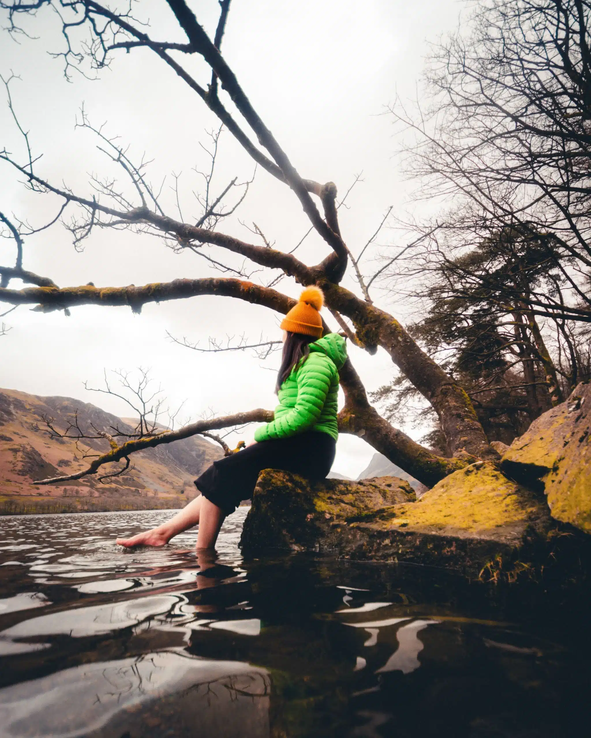 Person wearing a yellow beanie and green puffer jacket sitting on a mossy rock by a lake with bare feet dipping into the water beneath a large tree.