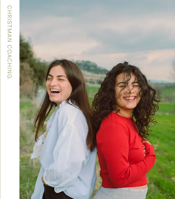 Two happy women standing back-to-back in a grassy field laughing, one with straight hair wearing a white shirt and the other with curly hair in a red top.