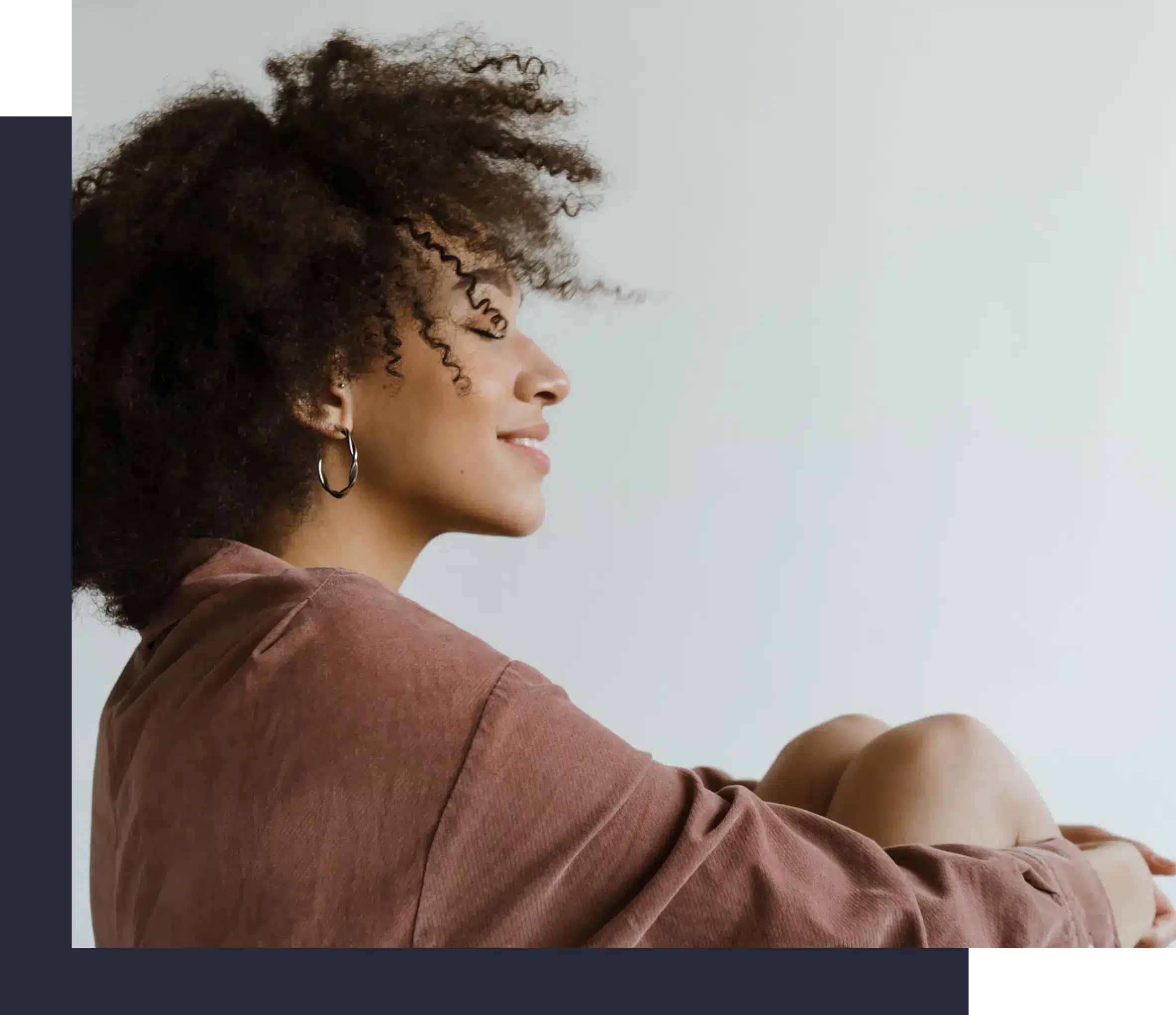 Side profile of a woman with natural curly hair and hoop earrings smiling peacefully with her eyes closed while hugging her knees.